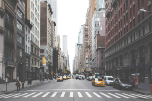 Eine belebte Stadtstraße mit hohen Gebäuden, fahrenden Fahrzeugen, Fußgängern auf dem Gehweg, Laternenpfählen und sichtbarem Himmel, mit Werbetafeln für New York City-Attraktionen im Hintergrund.