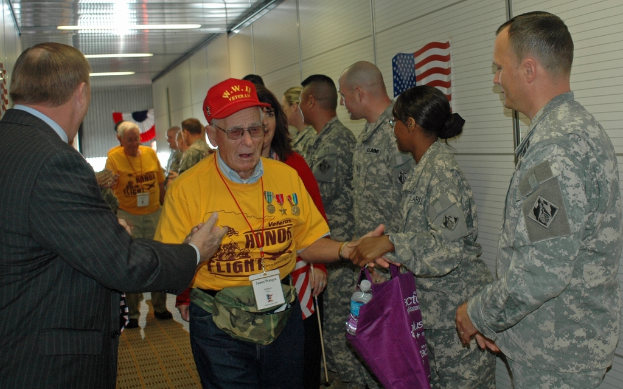 Eine Gruppe von Menschen und Soldaten steht in einem Raum mit einer Flagge an der rechten Wand und Lichtern an der Decke.