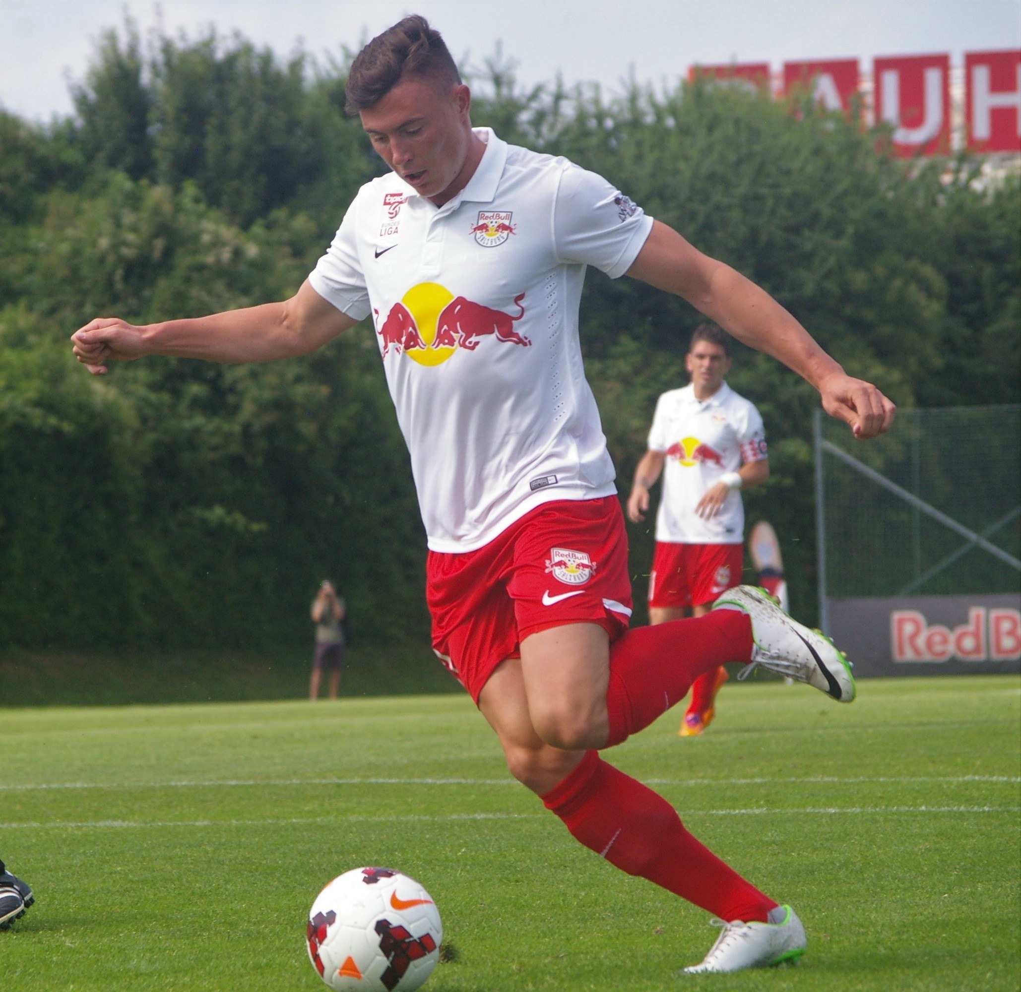 Ein professioneller Fußballspieler kickt einen Ball auf einem Rasenfeld mit Bäumen und einem klaren blauen Himmel im Hintergrund, in der Nähe einer Tafel mit der Aufschrift "RB Leipzig."