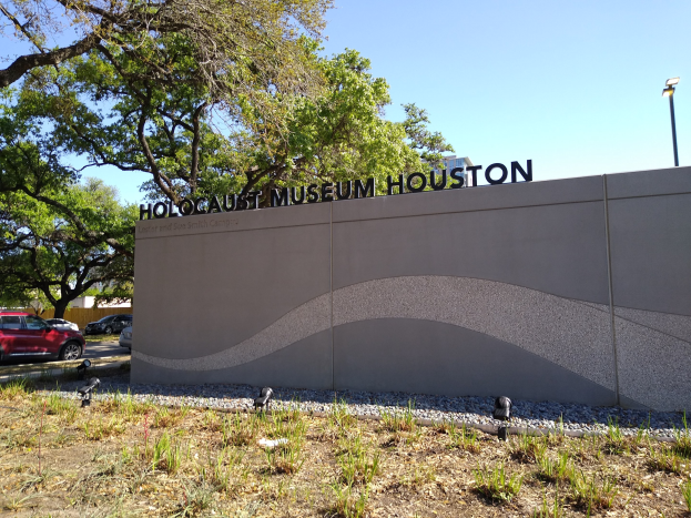 Außenansicht des Houston Holocaust Museums mit einer Gedenkwand, Steinen, Vegetation, Fahrzeugen, einer Straßenlaterne und einem bewölkten Himmel.