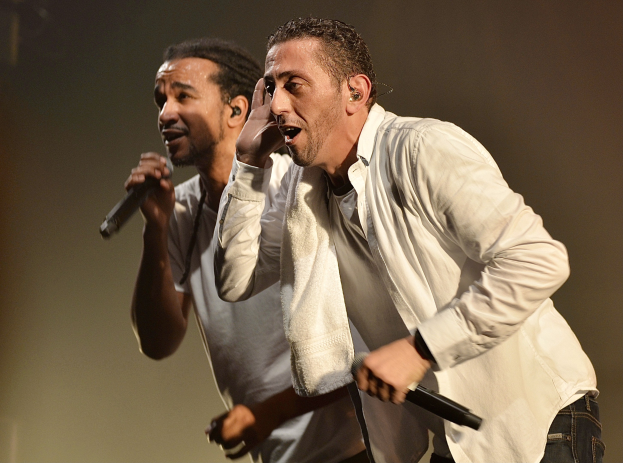 Zwei Männer in legerer Kleidung, die nebeneinander auf einer Bühne stehen und je ein Mikrofon halten.