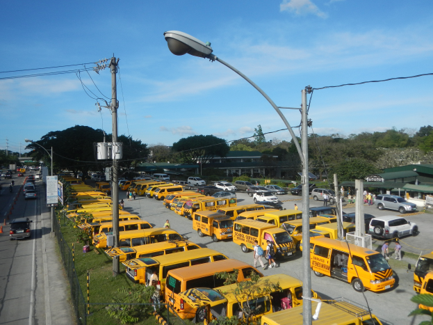 Eine große Anzahl gelber Schulbusse, die auf der Seite einer Straße geparkt sind, mit Menschen, die auf dem Gehweg gehen, Strommasten mit Drähten, Laternen, Bäume, Gebäude und ein bewölkter Himmel im Hintergrund.