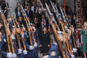 Gruppe von Menschen in militärischer Kleidung vor einer Menge stehend, einige halten Gewehre, mit Säulen und einer Pflanze im Hintergrund.