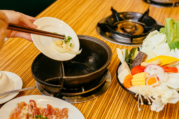 Eine Person hält einen Teller in der linken Ecke des Bildes, mit einem Tisch im Hintergrund, der einen Gasherd, eine Pfanne, eine Schale mit Obst und einen Teller mit Essen zeigt.