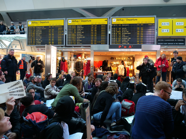 Große Gruppe von Menschen in einem Flughafen, einige mit Taschen und Papieren sitzend, andere stehend, mit Texttafeln, Schaufensterpuppen in Kleidern und Deckenbeleuchtung im Hintergrund, was auf eine Demonstration hindeutet.