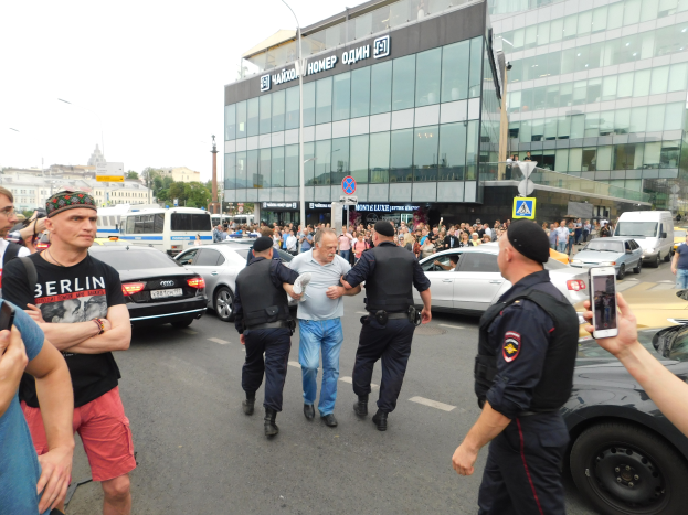 Eine Gruppe von Menschen, die auf einer Straße neben geparkten Autos gehen, einige halten Mobiltelefone, mit Gebäuden, Schildern, Bäumen und einem klaren blauen Himmel im Hintergrund, wahrscheinlich während einer Demonstration in Moskau mit Polizeipräsenz.