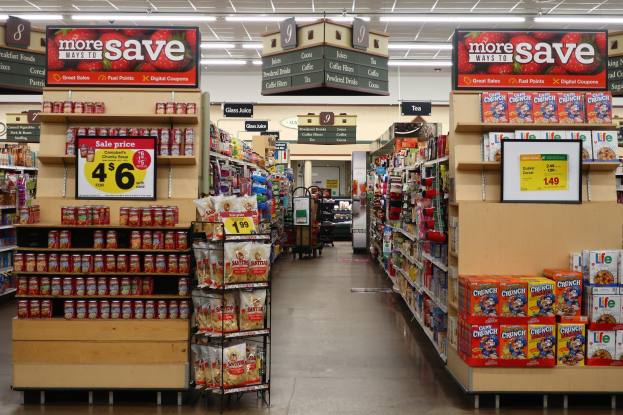 Ein Innenraum eines Lebensmittelladens mit Regalen und Boden voller verschiedener Lebensmittel, Deckenbeleuchtung und Informationsschilder im Hintergrund, mit einer Person in der Ferne sichtbar.