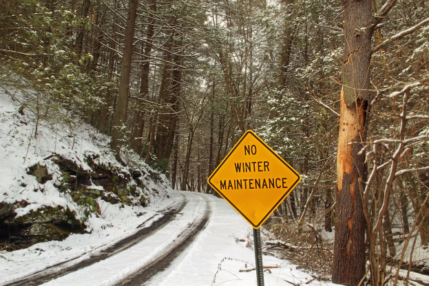 Kein Winterdienst-Schild an der Seite einer verschneiten Straße, umgeben von schneebedeckten Bäumen und einem klaren Himmel.