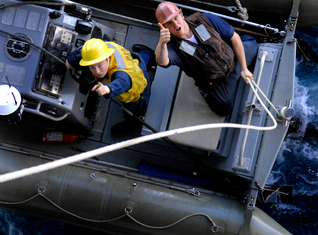 Zwei Männer in gelben Helmen und Sicherheitswesten auf einem Boot, einer hält ein Seil, scheinen eine Rettungsaktion im Wasser durchzuführen.