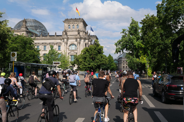 Eine Gruppe von Menschen, die auf Fahrrädern eine von Bäumen gesäumte Straße in Berlin, Deutschland, entlangfahren, mit Gebäuden auf der linken Seite und einer Bushaltestelle auf der rechten Seite unter einem bewölkten Himmel.