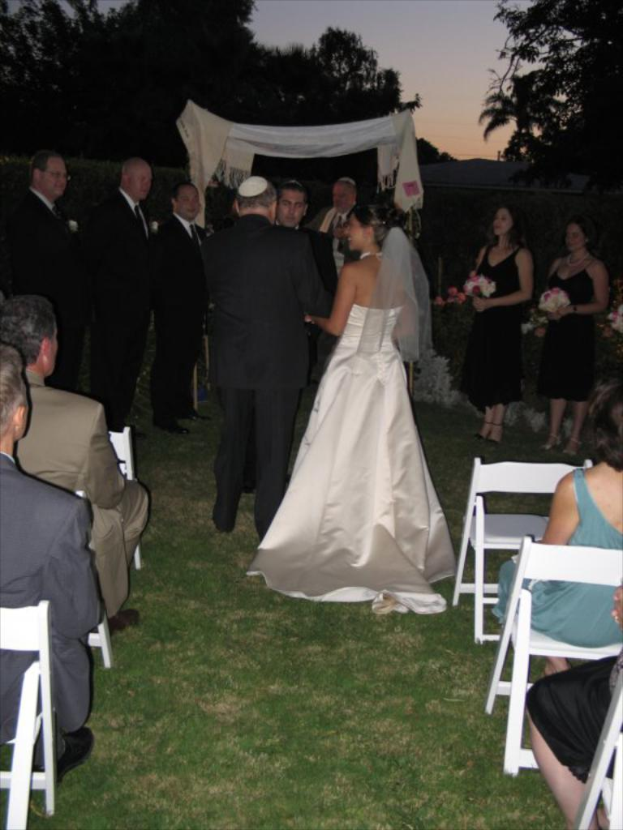 Eine Gruppe von Menschen bei einer Hochzeit, wobei Frauen auf der rechten Seite Blumensträuße halten, während andere auf Stühlen sitzen und Bäume im Hintergrund zu sehen sind.
