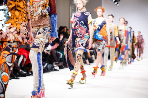 Eine Gruppe Von Frauen, Die Eine Laufsteg In London Fashion Week Heruntergehen, Mit Sitzenden Zuschauern Und Bühnenbeleuchtung Im Hintergrund.