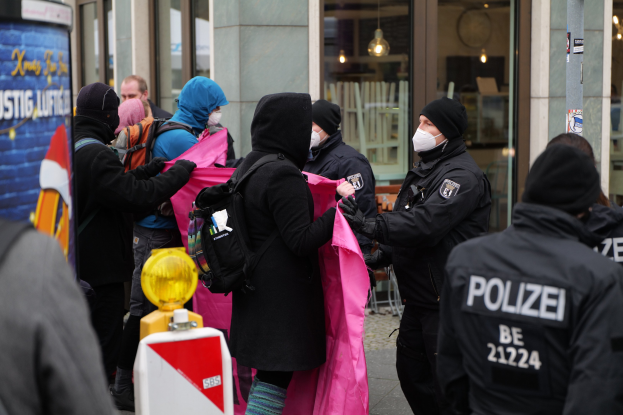 Eine Gruppe von Menschen steht vor einem gläsernen Gebäude mit Deckenbeleuchtung, einige tragen Mützen und tragen Taschen oder Kleidung, während ein Polizeibeamter eine Frau festnimmt; ein Pfahl mit Text und ein Wasserzeichen sind sichtbar.