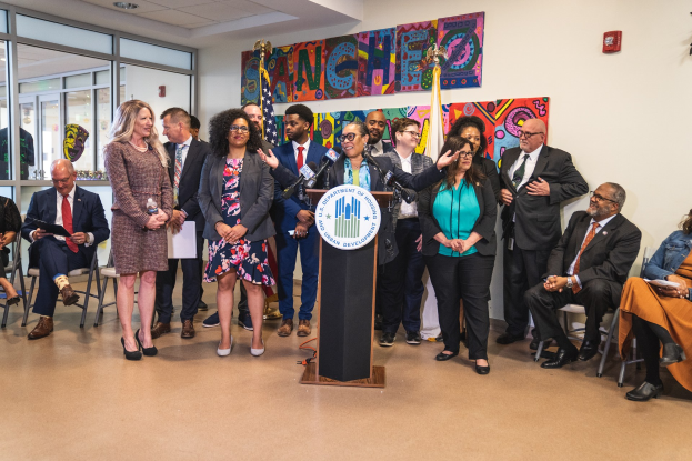 Eine Gruppe von Menschen steht vor einem Rednerpult mit einem Mikrofon und einer Tafel mit dem Text "Sancho Stadtverordneter tritt dem Stadtrat bei", während andere auf Stühlen sitzen oder in der Nähe stehen; Flaggen, ein Feuerlöscher und Deckenleuchten sind an der Rückwand zu sehen.