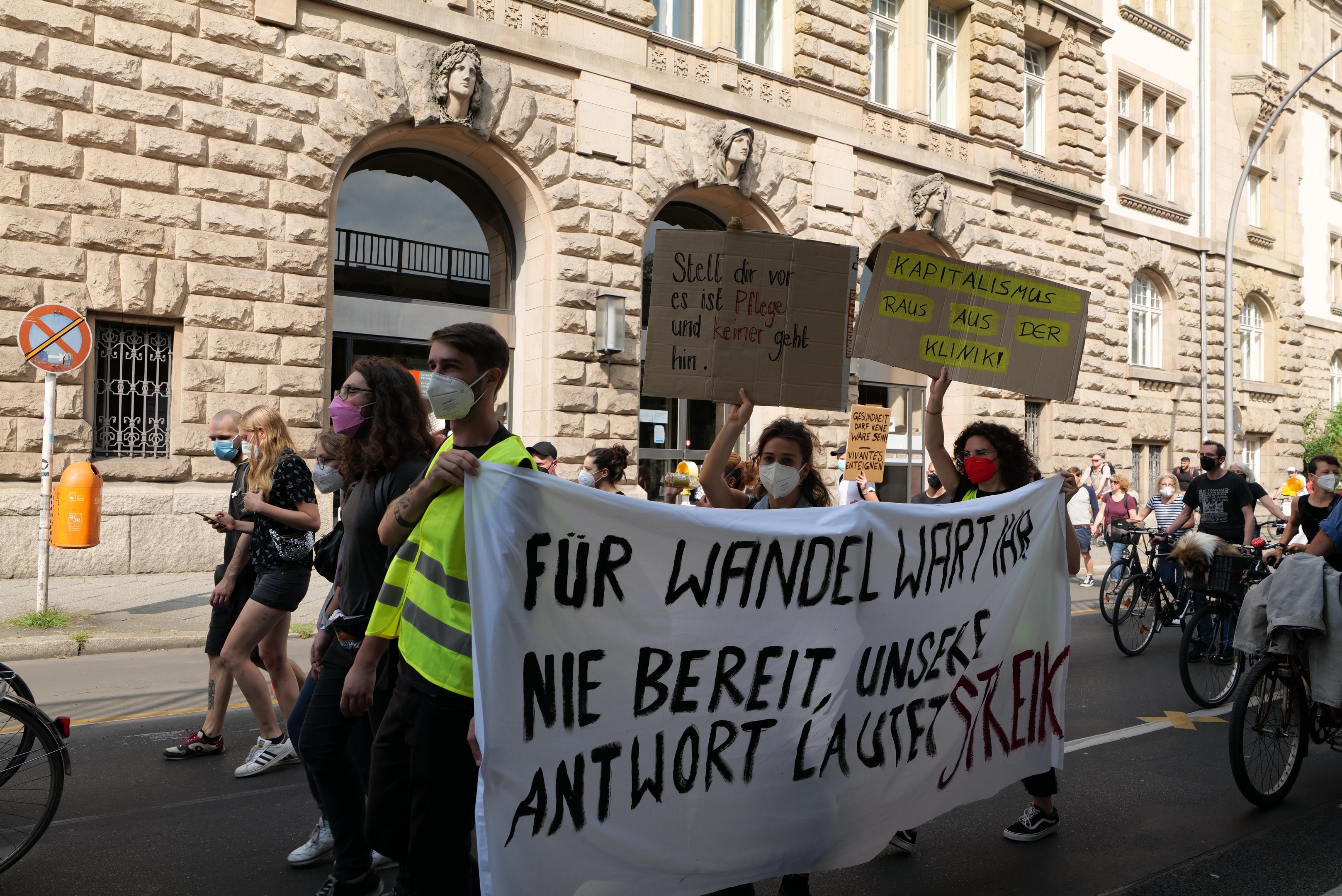 Eine Gruppe von Menschen marschiert auf einer Stadtstraße, einige halten Schilder und andere fahren Fahrräder, im Hintergrund ein historisches Gebäude mit Rundbogenfenstern, Säulen und Skulpturen.