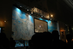 Eine Bühne mit einem Bildschirm in der Mitte, vor der eine Gruppe von Menschen sitzt.