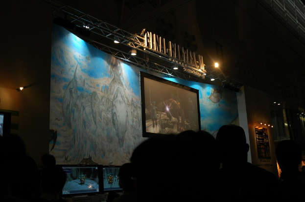 Eine Bühne mit einem Bildschirm in der Mitte, vor der eine Gruppe von Menschen sitzt.