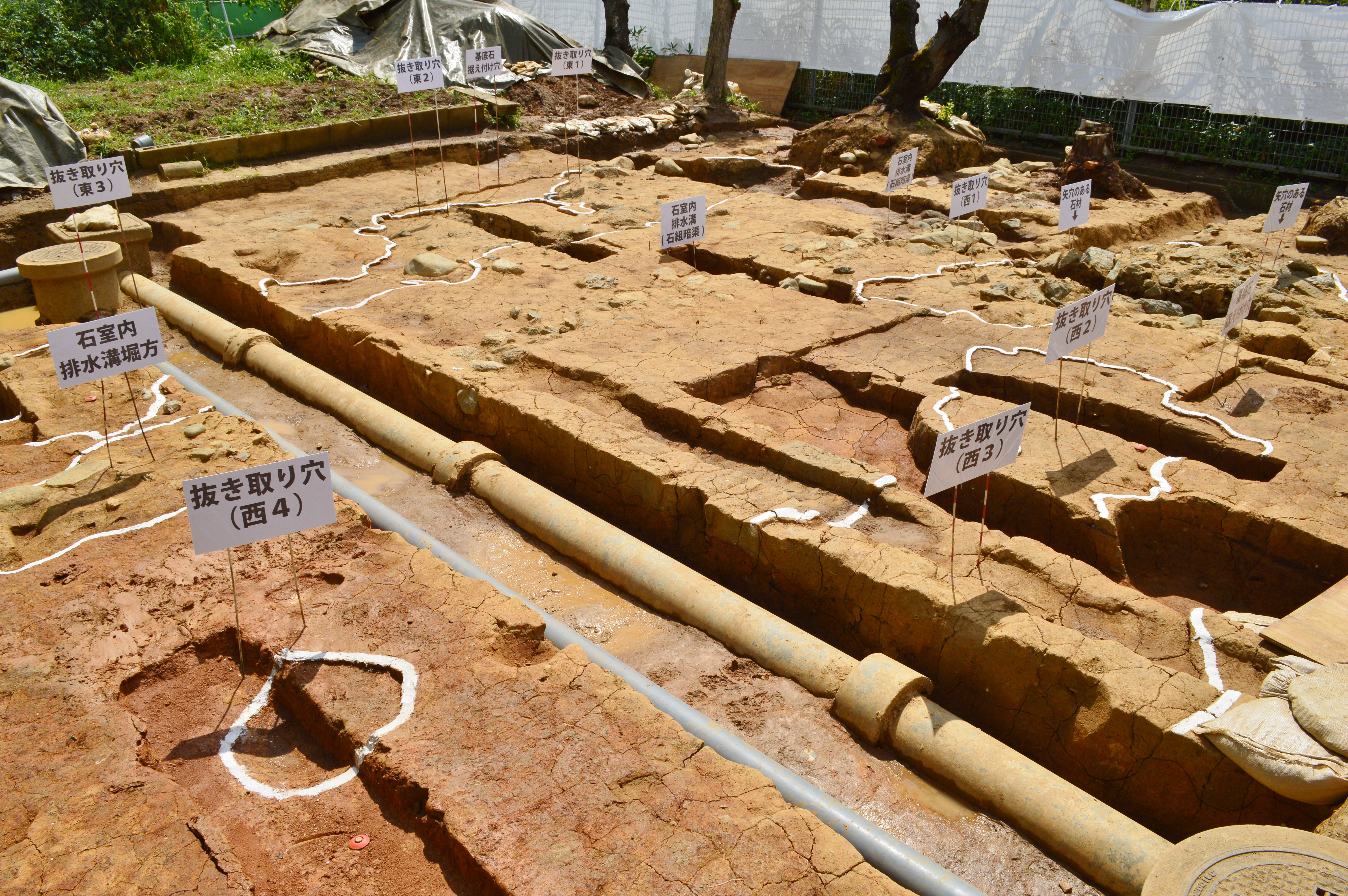 Große Erdaushubstelle mit Schmutz, umgeben von Bäumen, Pflanzen, Gras, einem Zaun, einer Abdeckung und einer Wand, mit Tafeln mit Text, Rohren und verstreuten Gegenständen.