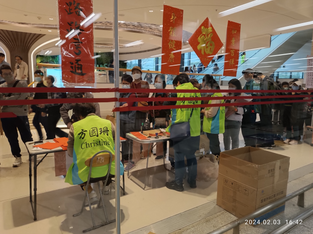 Eine Gruppe von Menschen in Gesichtsmasken steht an einem Tresen in einem Einkaufszentrum an, mit Stühlen, Tischen, Bannern und einem Geländer, sowie einem Karton und einem Wasserzeichen auf der rechten Seite.