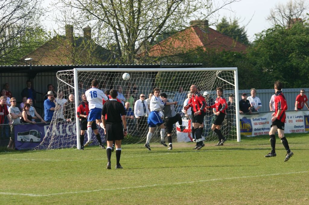 Fußballspieler spielen auf einem Feld mit einem Tor, während Zuschauer dahinter stehen; im Hintergrund sind Bäume und Häuser zu sehen.