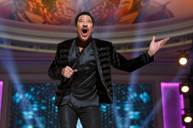 Lionel Richie performs bei den Grammys in einem schwarzen Kleid und hält ein Mikrofon in der Hand, mit einer beleuchteten B├╝hne und einem Bild im Hintergrund.