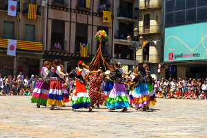 Eine Gruppe Frauen in bunten Kleidern tanzt auf der Straße, umgeben von einer Menschenmenge, mit Gebäuden, Geländern, Balkonen, Flaggen, einem Laternenmast und einer Tafel mit Schrift im Hintergrund.