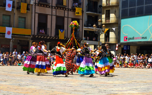 Eine Gruppe Frauen in bunten Kleidern tanzt auf der Straße, umgeben von einer Menschenmenge, mit Gebäuden, Geländern, Balkonen, Flaggen, einem Laternenmast und einer Tafel mit Schrift im Hintergrund.