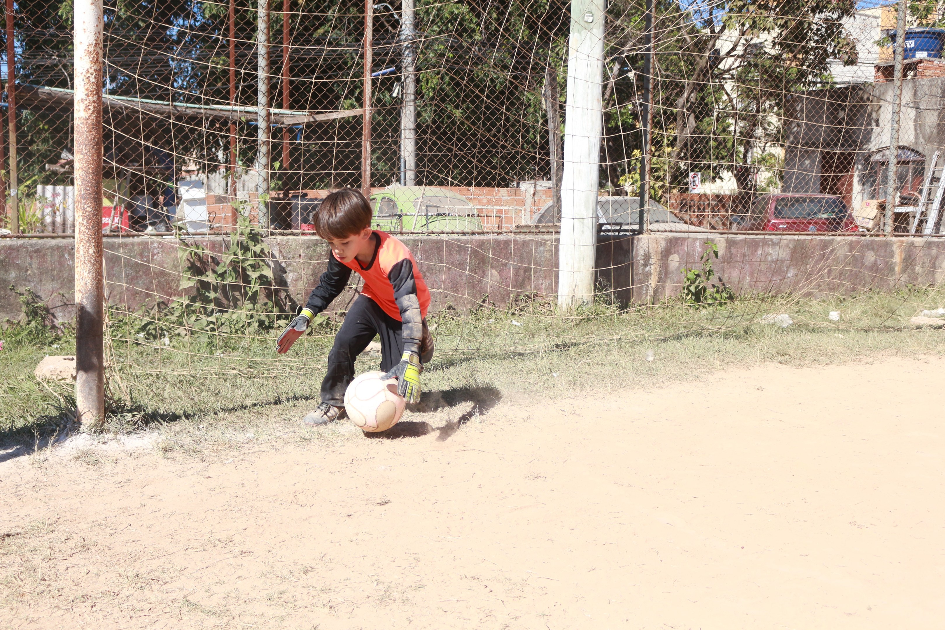 Ein junger Junge spielt Fußball auf einem staubigen Feld mit Gras, Pflanzen, Pfählen, einem Zaun, einer Wand, Bäumen, Fahrzeugen, Gebäuden und einem bewölkten Himmel, während er aktiv spielt und Schuhe trägt.