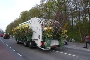 Ein Müllwagen mit Blumen auf der Ladefläche fährt eine Straße entlang, umgeben von Bäumen und einem klaren blauen Himmel, mit ein paar Menschen und Fahrrädern im Hintergrund.