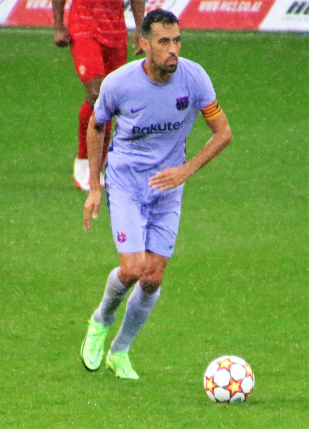 Ein Fussballspieler in einem Sportoutfit und -schuhen, der auf einem Feld mit einer textgefüllten Tafel im Hintergrund läuft.