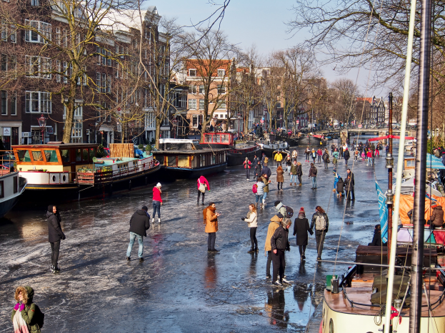 Eine Gruppe von Menschen, die auf einem Kanal in Amsterdam Eislaufen, umgeben von Booten, Bäumen, Gebäuden mit Fenstern, Laternenmasten und einem bewölkten Himmel.