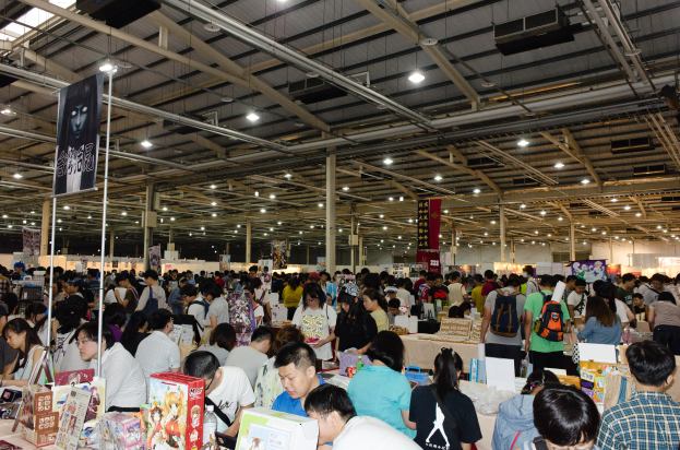 Eine große Gruppe von Menschen umringt Tische voller Bücher, Kisten und anderer Gegenstände auf einem Buchmesse, mit Bannern und Lichtern darüber.