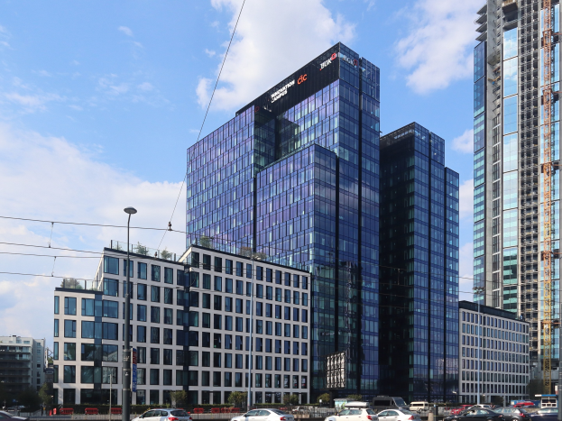 Außenansicht des Deutschen Bank-Hauptsitzes in Frankfurt, Deutschland, mit einem großen Gebäude mit vielen Fenstern, umgeben von städtischer Infrastruktur wie Straßenlaternen, Fahrzeugen, Grünflächen und einem bewölkten Himmel.
