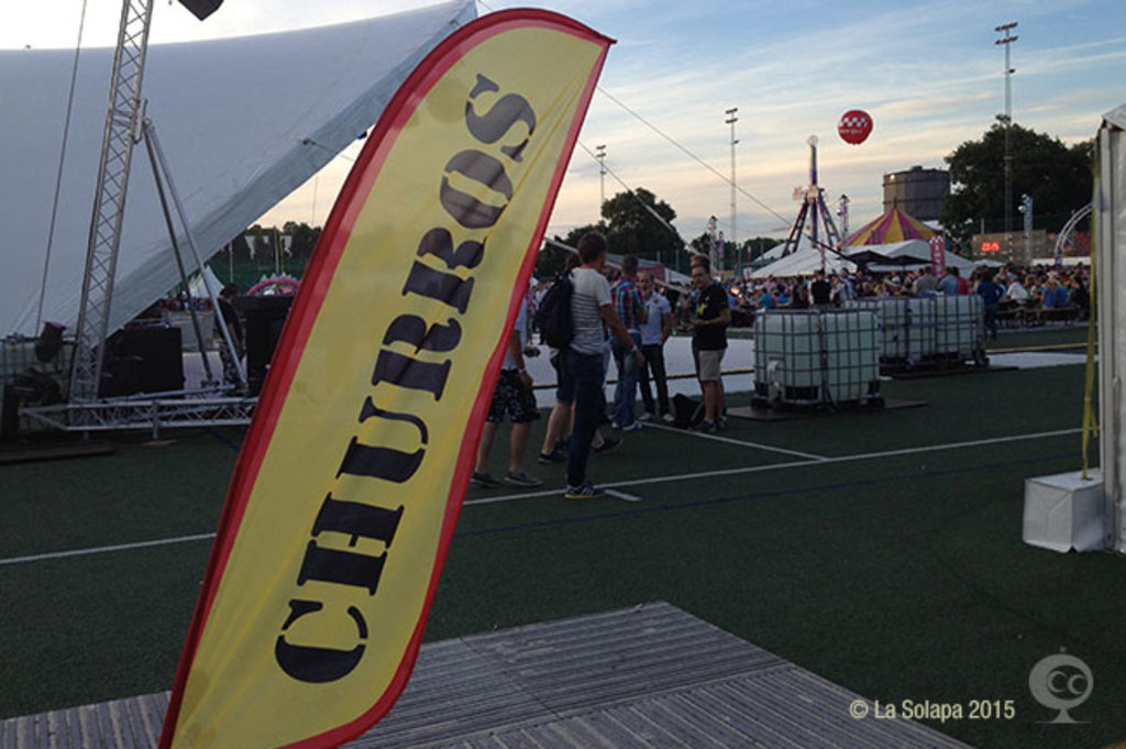 Eine lebhafte Karnevalsszene mit Zelten, einem zentralen Mast, Menschen, Bäumen, Strommästen, Straßenbahnen und einem eisernen Stand auf der linken Seite unter einem klaren Himmel.