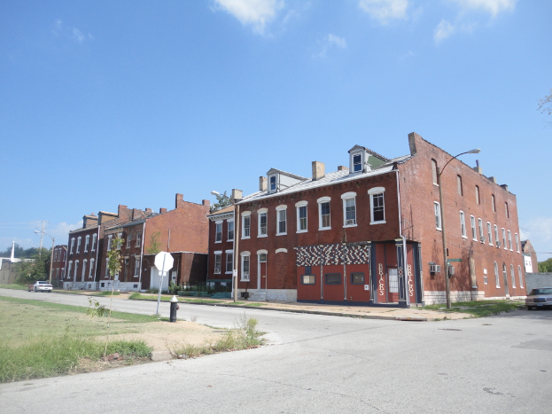 Ein Gebäude mit Pfählen und Straßenlaternen, umgeben von Bäumen und Fahrzeugen auf beiden Seiten, mit einem Weg, Gras und einem Feuerlöscher unten und dem Himmel oben.